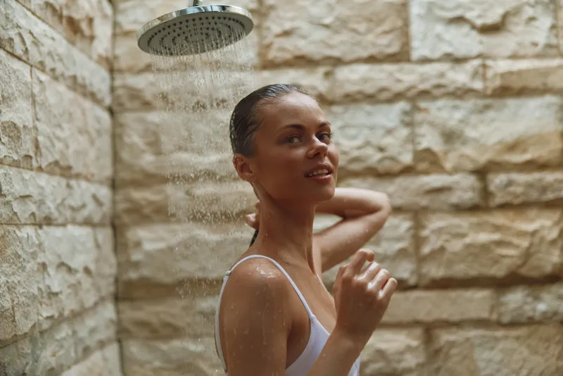 young woman enjoying outdoor shower with natural stone wall background, relaxed mood, wet skin, casual white swimsuit under soft natural light