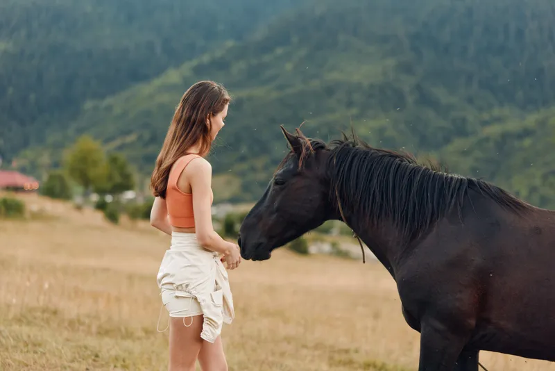 A young woman stands in a rural field, gently facing a horse as they share a quiet moment. The scene conveys calm, connection, and a serene, natural ambiance.