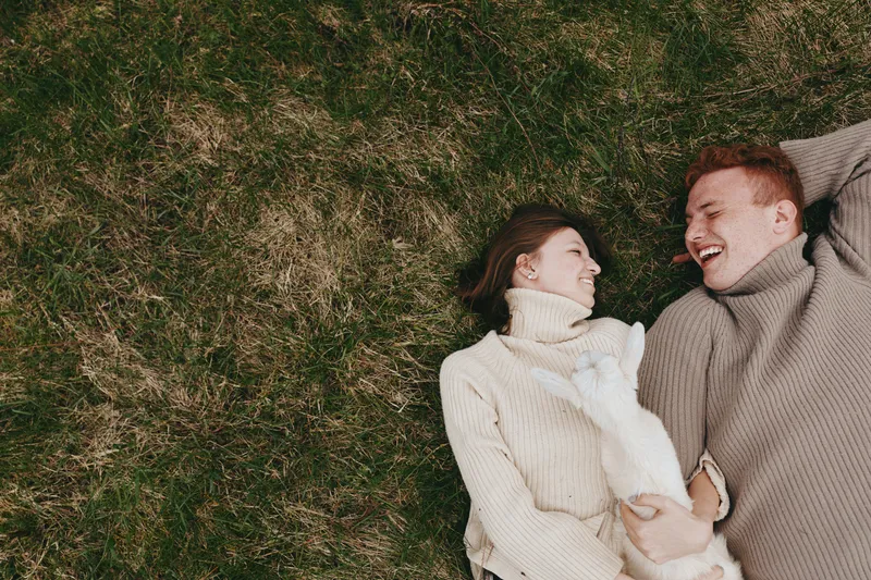 Young couple lying on grass in warm sweaters laugh and enjoy a cozy moment together holding a small white baby goat in a natural outdoor setting during autumn.