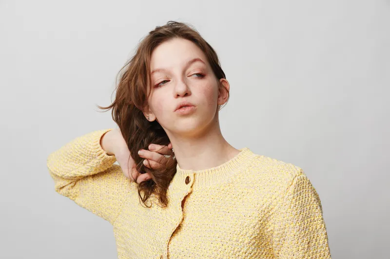 Young woman in yellow knitted sweater looks away thoughtfully with hand behind head on white background. Concept of contemplation, casual style, and natural beauty.