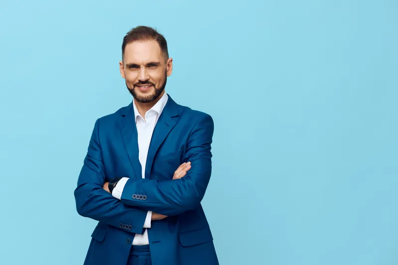 A professional businessman stands with arms crossed against a solid blue background, conveying confidence, poise, clarity, focus, and calm emotion in a clean studio portrait.