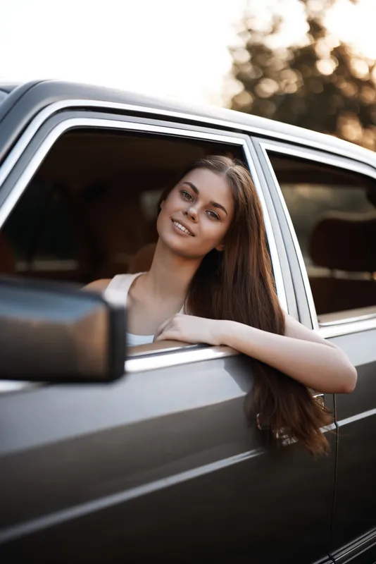 Young woman smiling in a car window during sunset, showcasing carefree emotions and a casual style, perfect for concepts of freedom and adventure.
