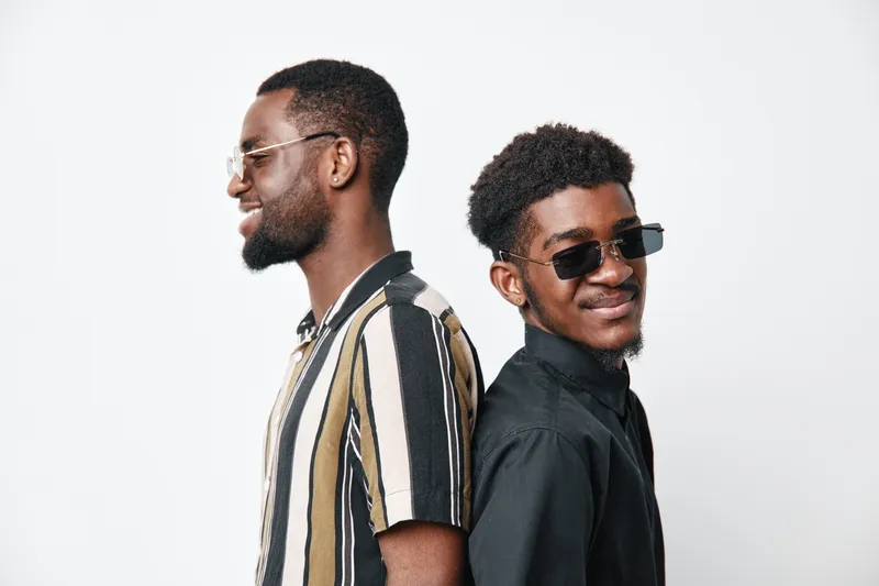 Two African American men with dark skin standing back to back wearing sunglasses and casual shirts, smiling and posing against white background in studio portrait.