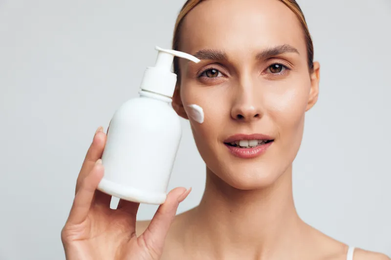 Young woman with clean face holds white bottle with cream on cheek, demonstrates skincare routine, natural beauty concept, isolated on light gray background closeup portrait.