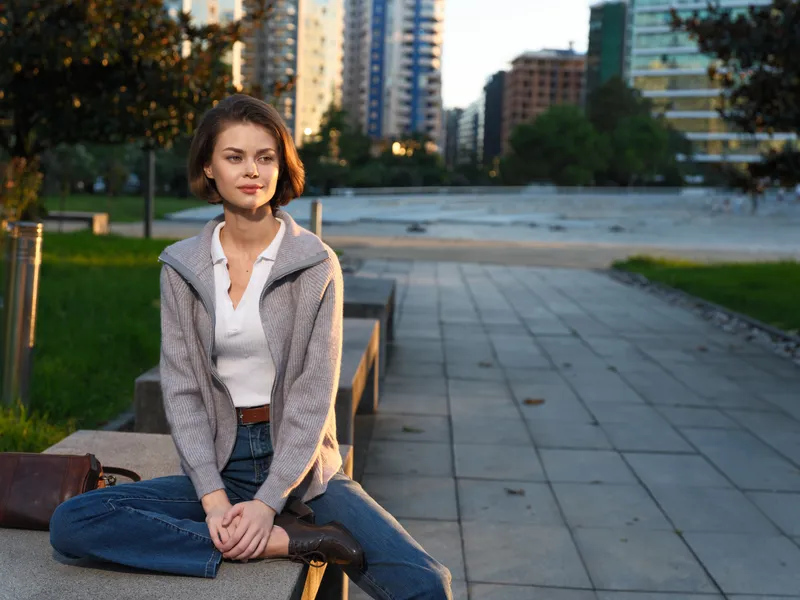 A stylish young woman sits on a concrete bench in an urban park, wearing casual attire. She gazes forward with a calm smile, exuding a relaxed, confident mood in a modern city setting.