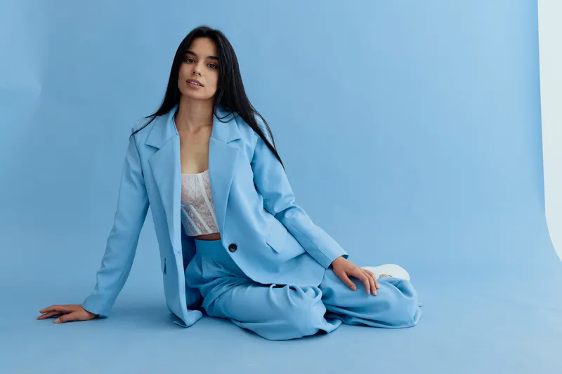 young woman in blue suit sitting on pale blue background, relaxed pose, long dark hair, modern casual fashion, soft natural lighting.