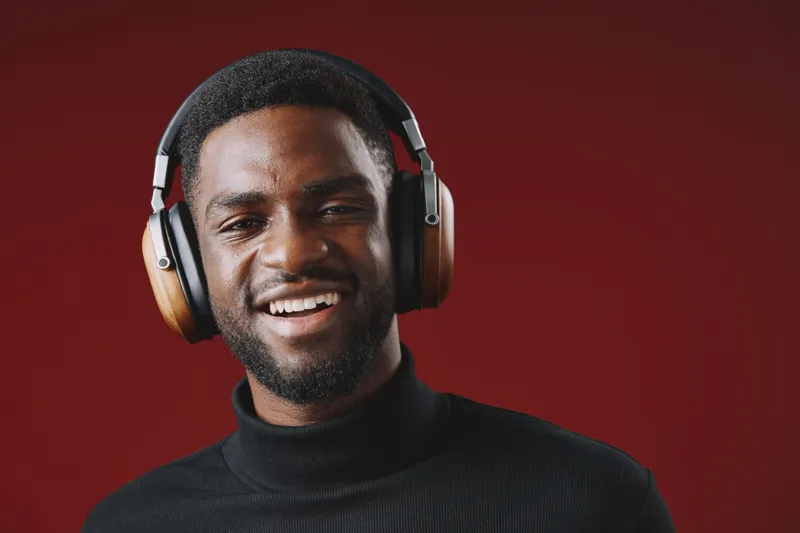Happy African man wearing headphones smiling against a deep red background, showcasing modern technology and joyful emotions in a stylish attire