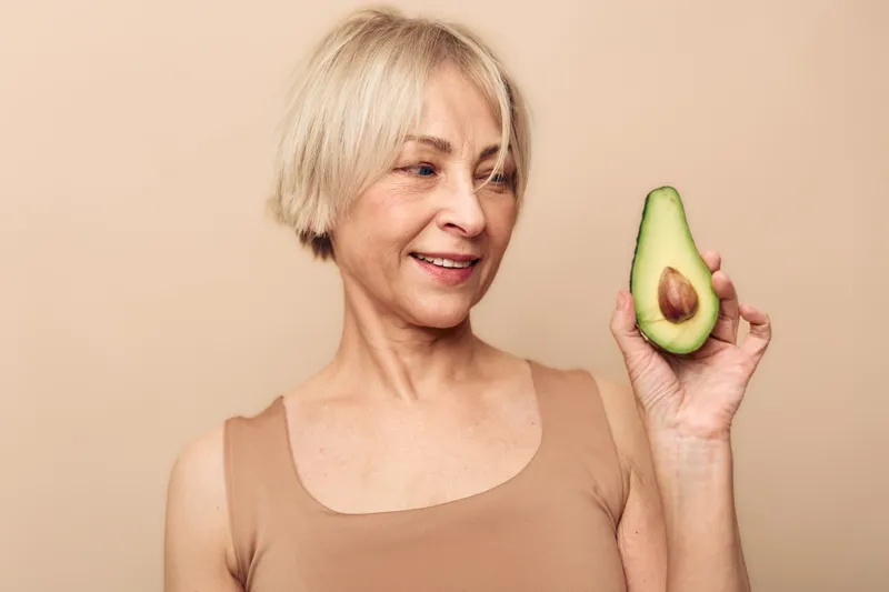 Smiling mature woman with short blonde hair holds half avocado close to face on beige background. Concept of healthy lifestyle, nutrition, natural food, and wellbeing for seniors.
