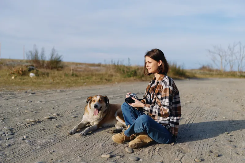 Woman sits on a dirt ground with a friendly dog beside her, outdoors in a barren landscape. She holds a small camera, enjoying quiet moments and companionship in wide open space.