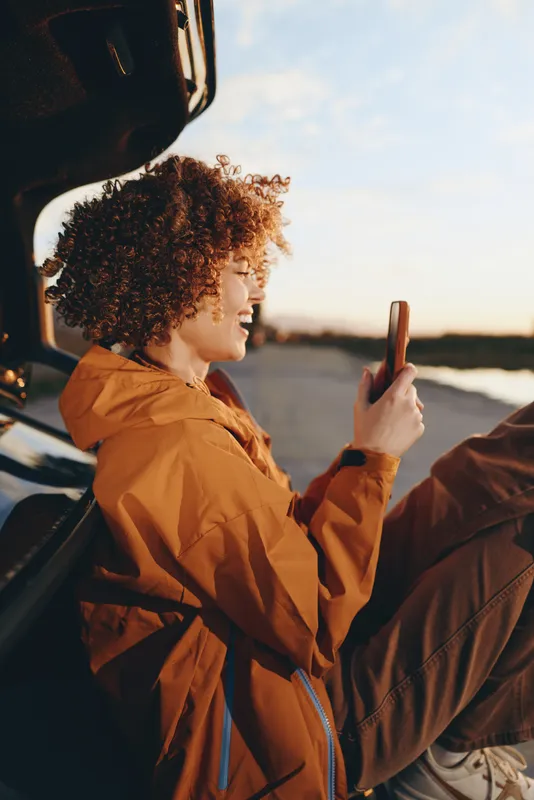 Lifestyle woman by a car at sunset, curly hair woman holds a smartphone for selfie, warm rim light on her face, outdoor travel moment, casual jacket, road atmosphere today peaceful