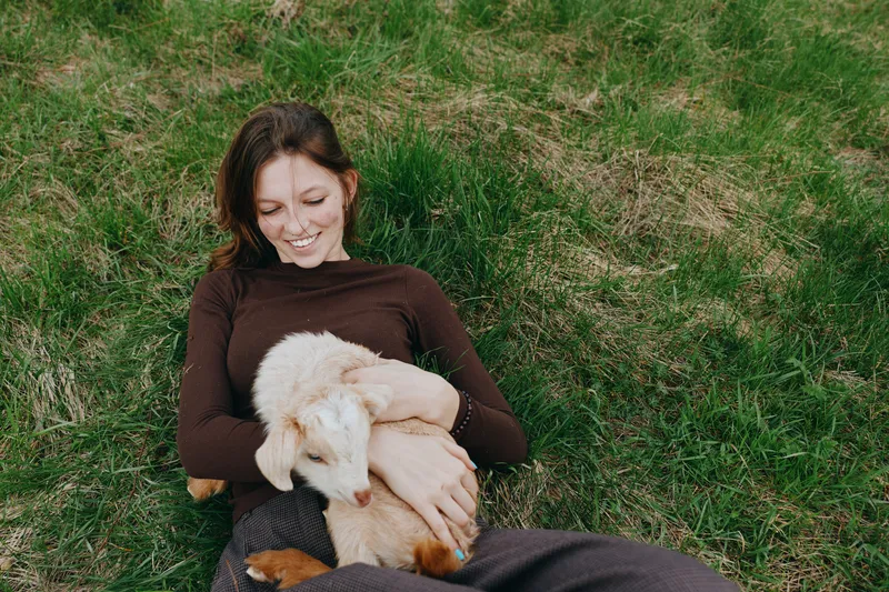 Cheerful woman outdoors, smiling while cuddling puppy on grass, sunlight lighting the scene, relaxed posture, natural colors, joyful lifestyle moment