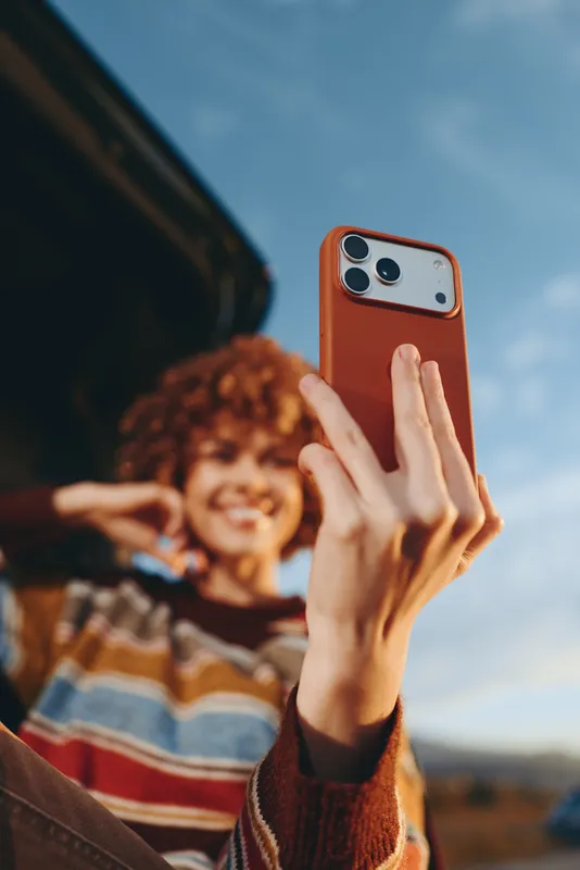 Woman selfie smartphone outdoor lifestyle smiling sweater sky hands portrait, holding a red phone case for a candid photo, sunny mountains, clouds and warm daylight.