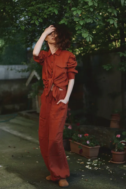 A woman in a red jumpsuit stands in a garden, posing thoughtfully with wind in her hair. An outdoor lifestyle moment highlighting casual fashion and natural light in a calm mood
