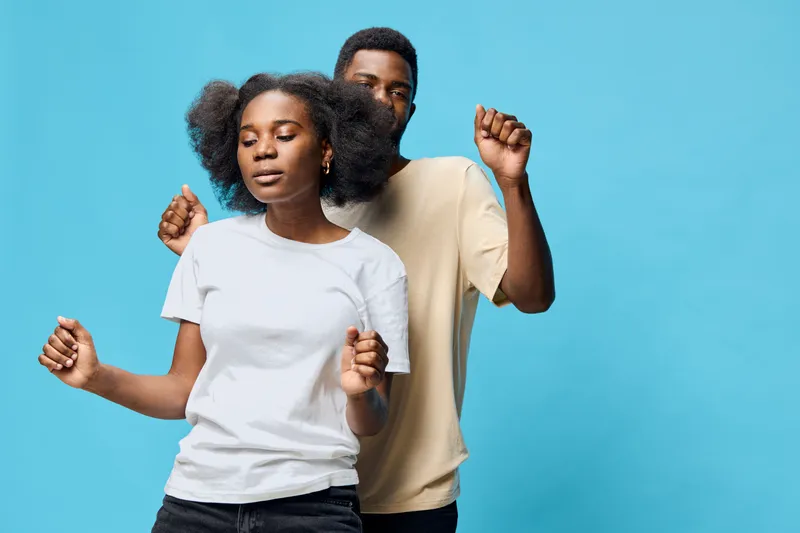 A joyful couple dancing together against a soft blue background, showcasing playful emotions and contemporary casual fashion in a vibrant setting