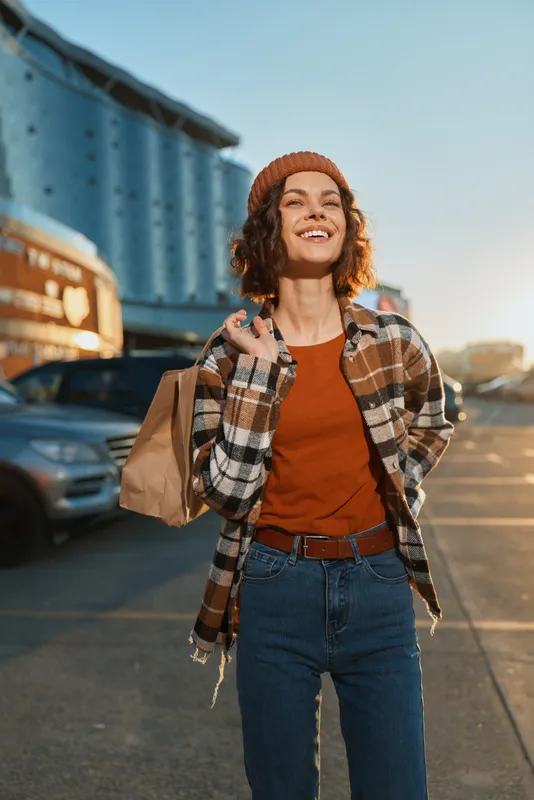 Woman smiling with shopping bag in urban parking lot at golden hour, candid lifestyle portrait showing authenticity, mindful living and emotional storytelling in warm glow.