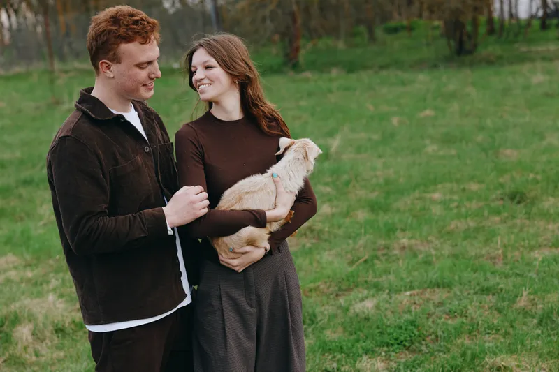 Young couple enjoys a peaceful moment outdoors as the woman gently holds a small goat while they smile and connect in a green natural field during a casual lifestyle shoot.