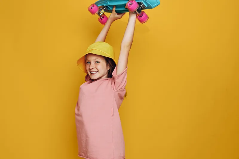 Happy girl in yellow hat holding blue skateboard above head smiling against yellow background. Playful and energetic youth concept.