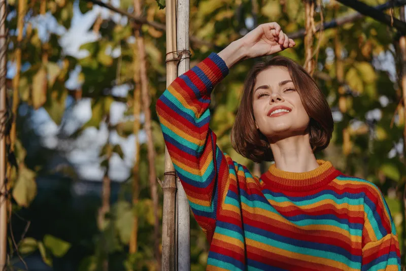 Woman in a rainbow sweater smiling outdoors, short hair bob, leaning on a bamboo pole in a sunny garden. Casual outfit, joyful 25-year-old model conveying relaxed travel and lifestyle vibe.