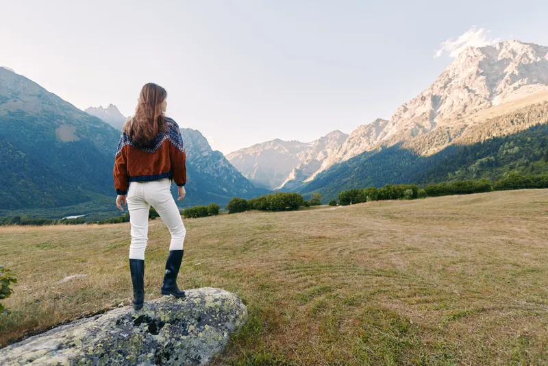 Woman stands on a rock in a mountain meadow wearing hiking boots, looking at panoramic mountains and valley. Outdoor nature view, travel adventure and serene alpine landscape solitude.