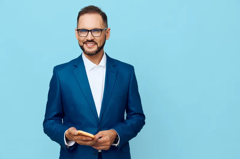 A confident business man in a blue suit stands against a solid blue background, projecting a friendly, professional vibe while holding a phone.