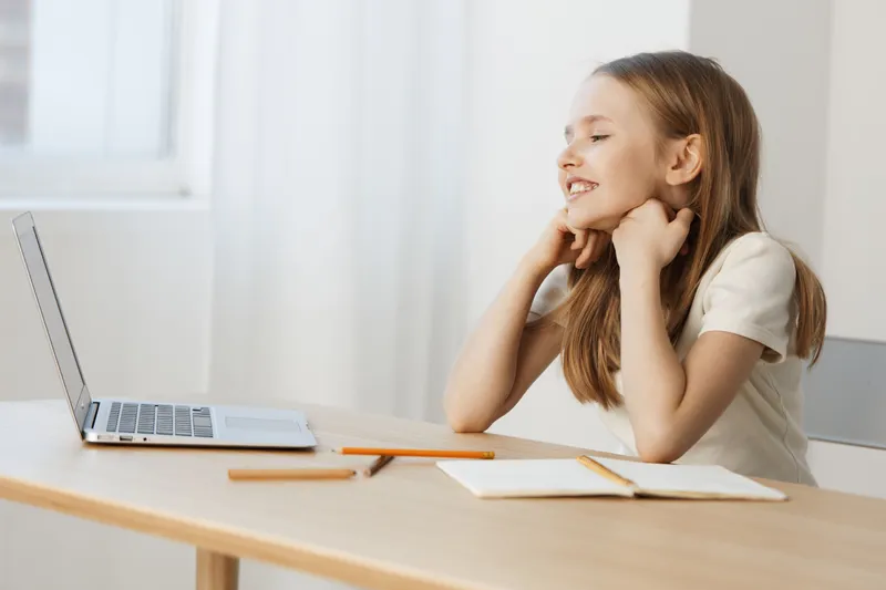 Happy girl enjoying online learning at home, smiling with excitement while using a laptop, surrounded by school supplies in a bright, minimalistic room