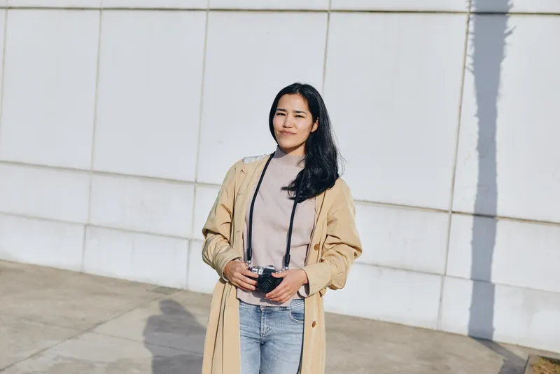 Casual woman standing outdoors in a beige coat, holding a phone, relaxed pose near a white wall, showcasing contemporary street style and everyday fashion.