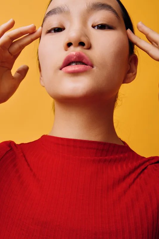Young woman with clear skin wearing red sweater posing with hands near face on orange background. Beauty, skincare, portrait, confidence, fashion and casual style in studio photo.