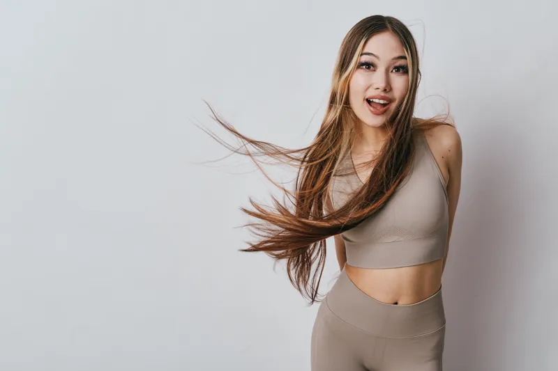 Young woman in sportswear with long hair and surprised face posing in studio. Fitness, healthy lifestyle, energetic female model with excitement and movement on grey background.