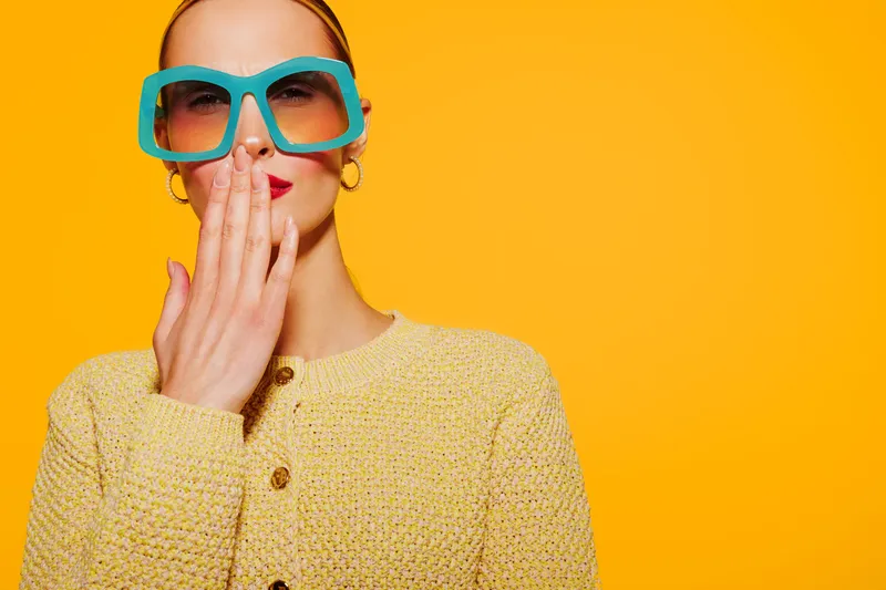Stylish woman in blue oversized sunglasses and yellow sweater covers mouth with hand on bright orange background. Concept for fashion, surprise, and modern style.