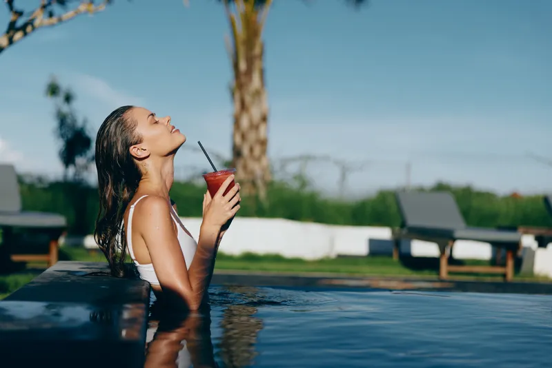 A woman relaxes in a sunny pool, sipping a red drink, eyes closed, savoring the warm breeze. A lifestyle moment of leisure, outdoor pleasure, and carefree refreshment.