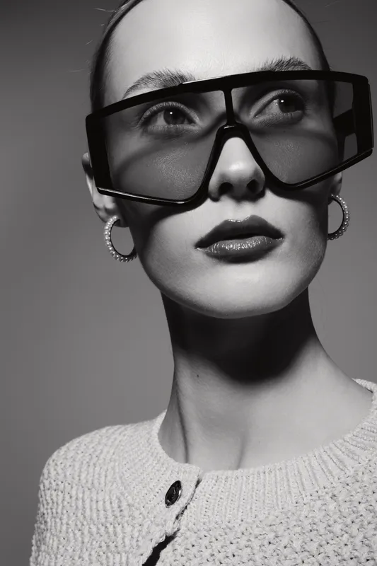 Portrait of a fair skinned Caucasian woman wearing large sunglasses and hoop earrings. She wears a knitted top and looks away in a stylish black and white fashion concept.