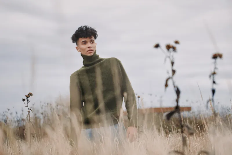 Young man in a green turtleneck stands among tall dry grasses under soft overcast light, photographed candidly to convey authenticity and a trustworthy, natural portrait feeling.