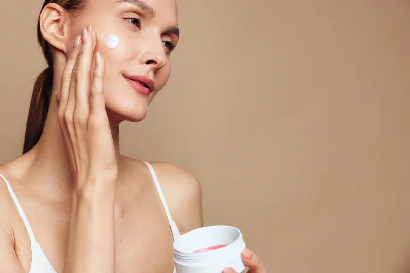 Young woman applies moisturizing cream on face with fingers holds jar of cosmetic product skin care routine on beige background natural beauty and healthy lifestyle concept.