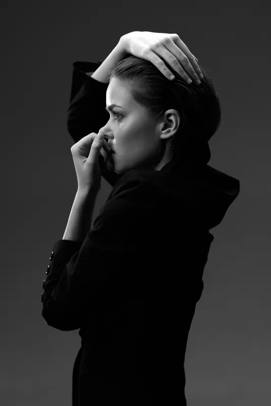 Black and white portrait of a thoughtful woman in a dark outfit, showing introspection and elegance in a minimalistic studio setting.