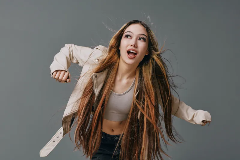 Young woman with long hair dancing and looking up with joyful expression on gray background. Casual outfit, movement and energy in studio with happy female model.