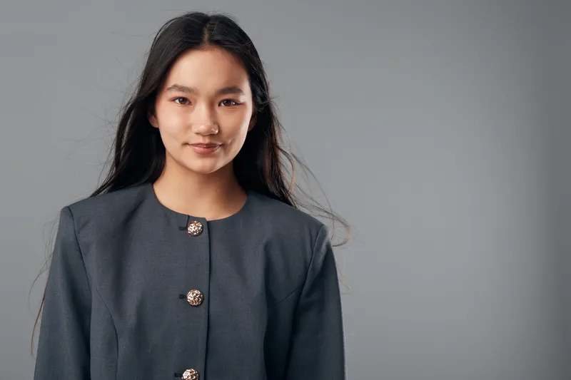 Young woman portrait with long dark hair and gray buttoned jacket posing on gray background. Confident female looking at camera with natural expression and relaxed pose in studio light.