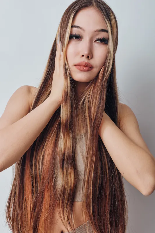 Woman with long brown hair posing with hands near face, looking at camera with neutral expression. Beauty portrait with natural makeup and smooth skin on gray background.