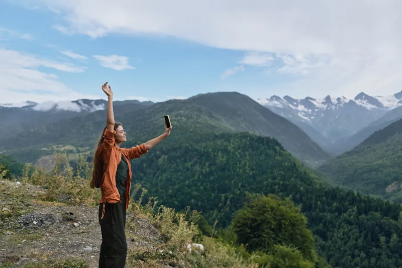 Woman selfie in an outdoor landscape among towering mountains, capturing nature and adventure in a bright scenic moment. Her pose expresses freedom and curiosity as clouds drift above alpine peaks.