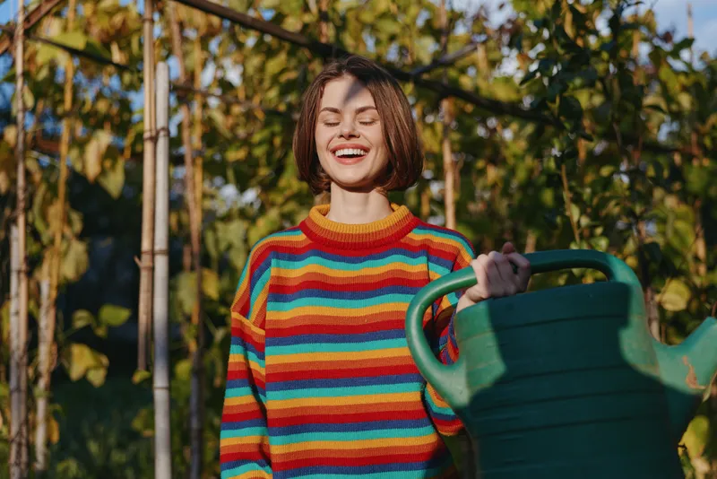 Woman with watering can in striped sweater in a sunlit garden, smiling young adult tending vine plants by trellis, outdoor lifestyle portrait showing joyful expression and casual outfit.