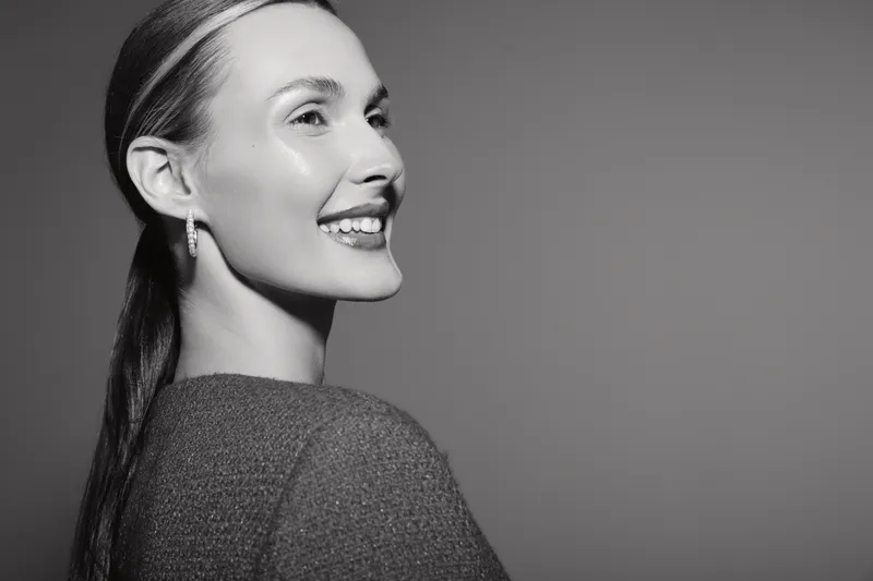 Smiling young Caucasian woman with fair skin and long hair looks sideways, wearing knitwear. Studio portrait in black and white with soft lighting.