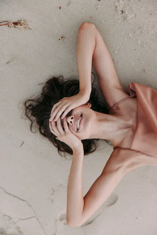 A relaxed woman lies on sandy beach wearing a soft pink dress, smiling softly as one arm rests over her face, capturing a warm, carefree moment in natural light.