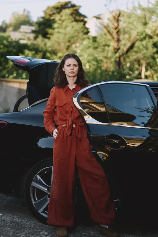 Confident woman in a rust red jumpsuit stands beside a sleek car outdoors, sunlit moment, casual chic vibe, relaxed posture, natural colors, lush background