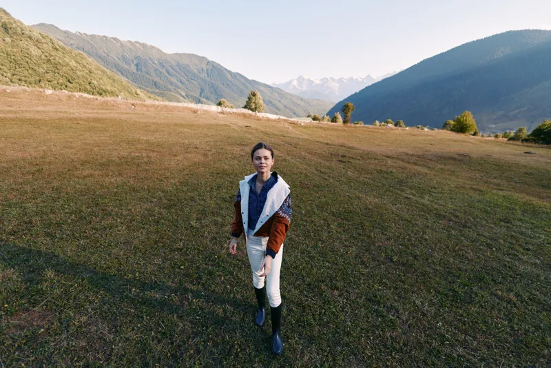Woman walking in a meadow with mountains and expansive landscape, nature portrait of a lone hiker in open field wearing jacket and boots during clear day with golden grass and distant peaks at sunrise