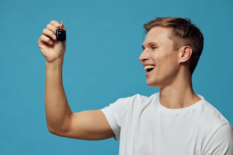 Smiling young man with short hair wearing a plain white t-shirt holding a car key up towards the camera isolated on a solid blue background studio portrait. People lifestyle concept with excitement an