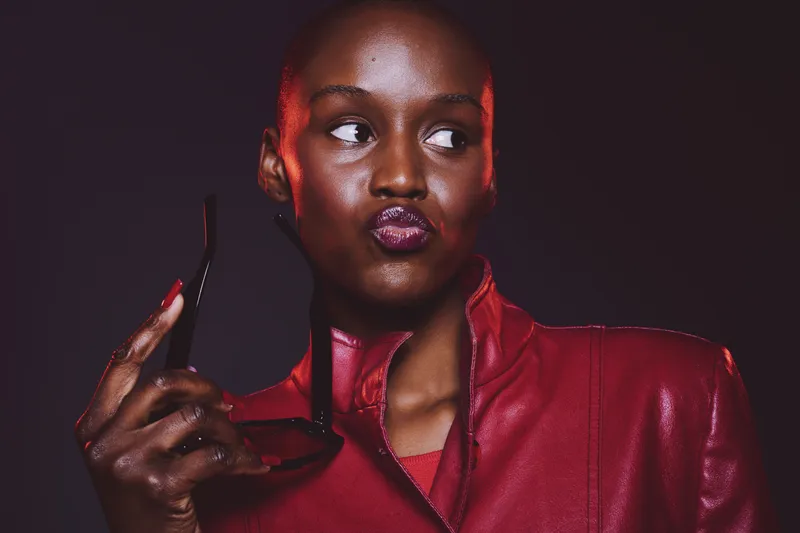 Portrait of confident black African woman with dark skin wearing red leather jacket holding sunglasses. Studio shot with dramatic lighting and expressive face looking sideways. Fashion and style
