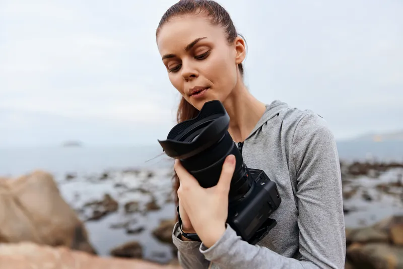 A thoughtful young woman holding a camera on a rocky beach, embodying creativity and passion for photography in a serene coastal environment