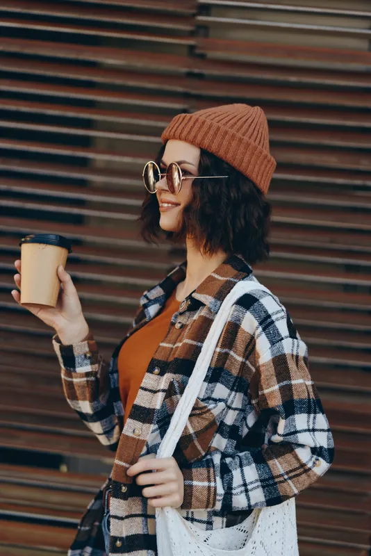 woman coffee beanie smile plaid tote glasses urban portrait of a young woman holding a takeaway cup, authenticity and candid lifestyle captured with golden hour glow, mindful living and emotional