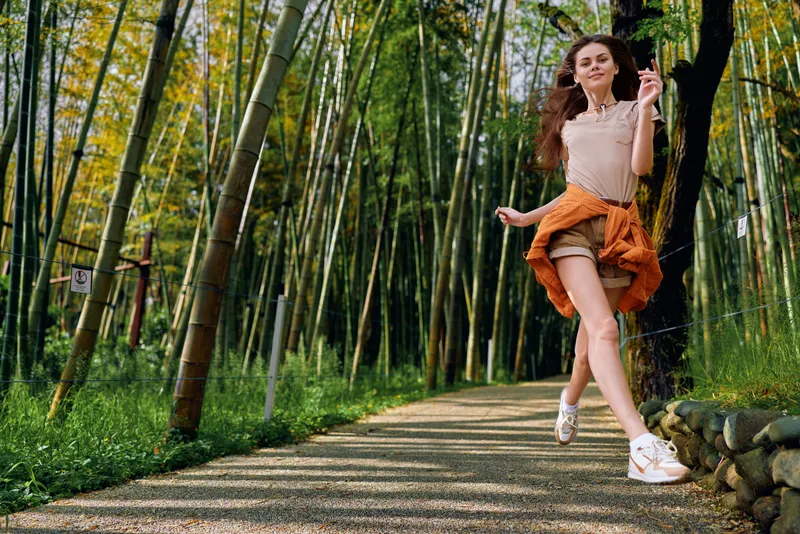 Woman jump along a bamboo path in a forest, casual summer outfit and smile, motion captured midair on a sunny outdoor walkway conveying joyful energy and carefree lifestyle.