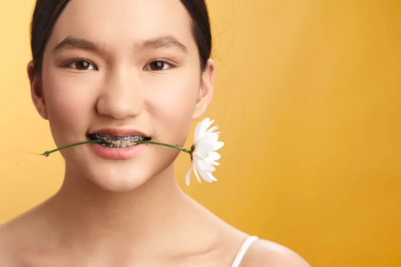 Asian woman with a flower between her teeth smiles softly in a warm yellow studio, presenting a playful, fresh portrait with natural charm and bright mood.