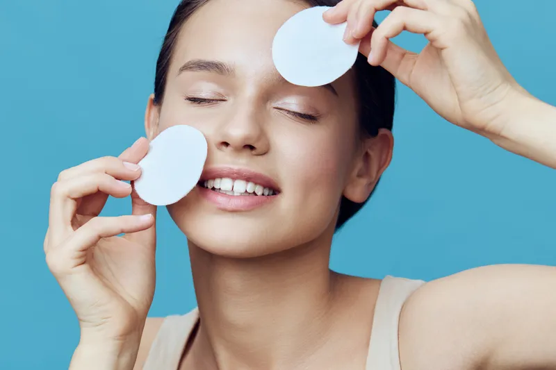 Young woman enjoying skincare routine holds cotton pads on her face, smiling against a bright blue background, exuding happiness and self care